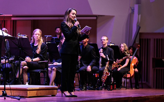 Esther spreekt het publiek toe op een podium, met een orkest achter zich